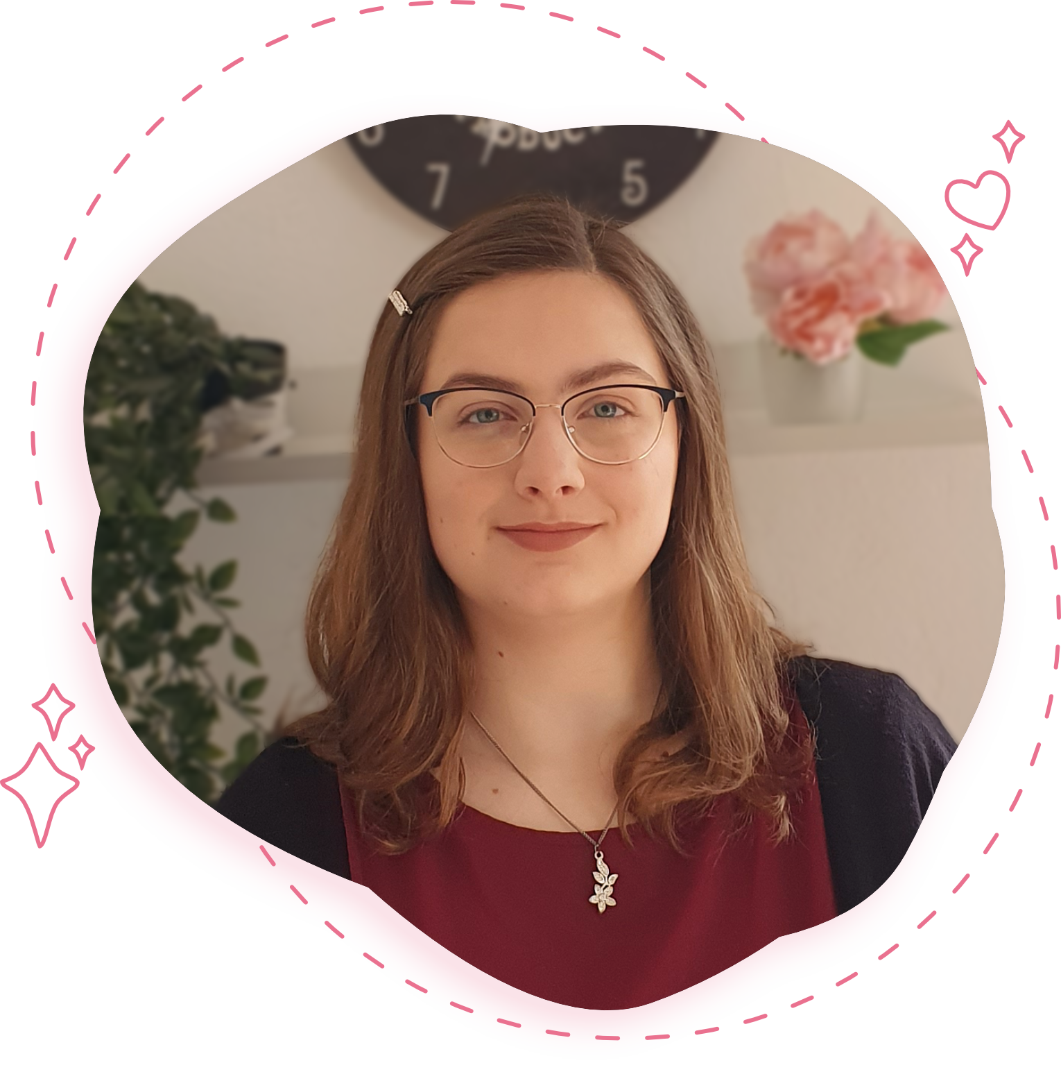 Laura Radetzky wearing a red shirt with a black cardigan. She wears a silver necklace with a flower charm. She has long brown hair and is wearing glasses. Her hair is down with a side part and she wears a silver hair pin. In the background, there are some flowers and a hanging ivy plant. In the top middle, there is a black clock.